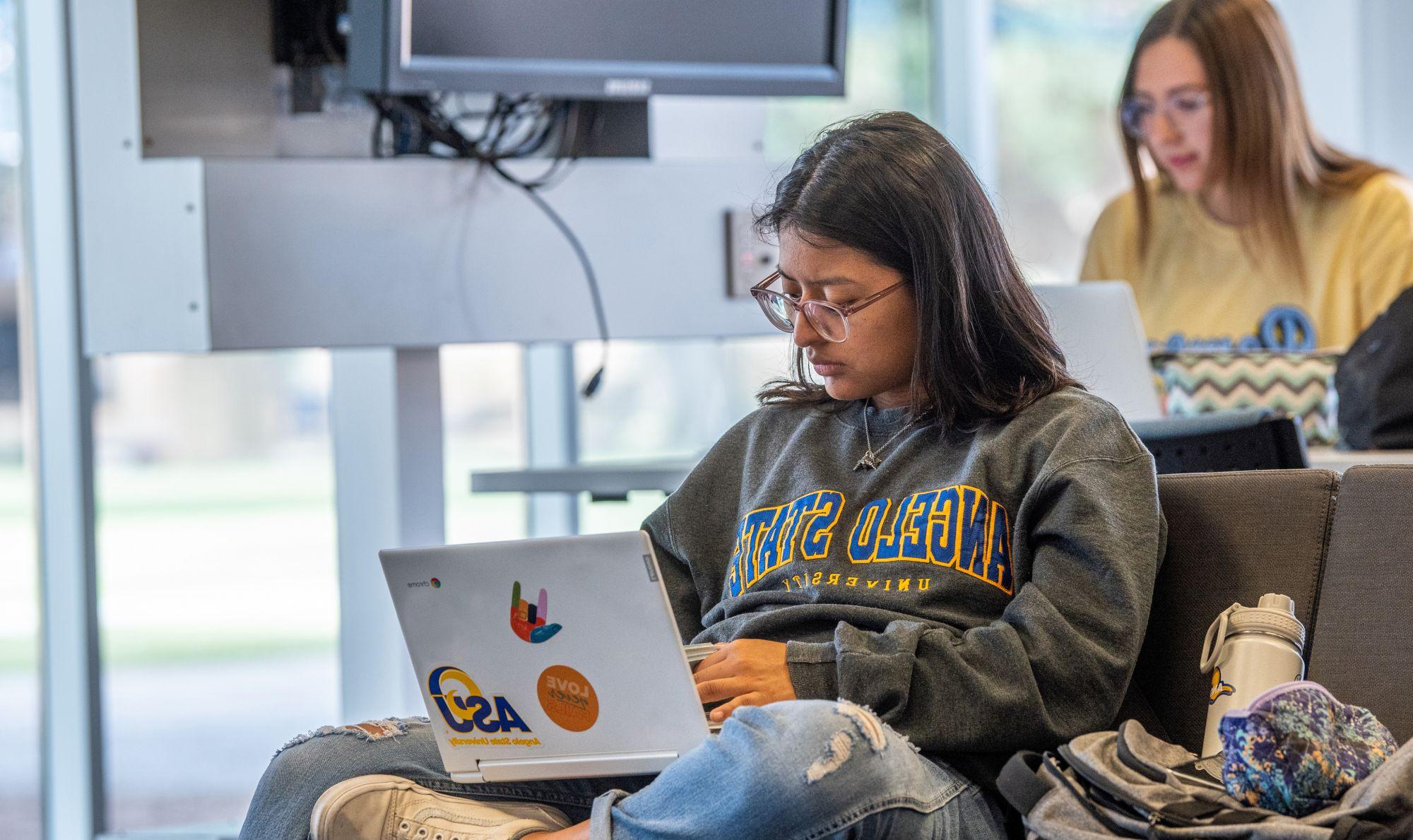 一个学生坐在图书馆用笔记本电脑学习，另一个学生在后台学习.