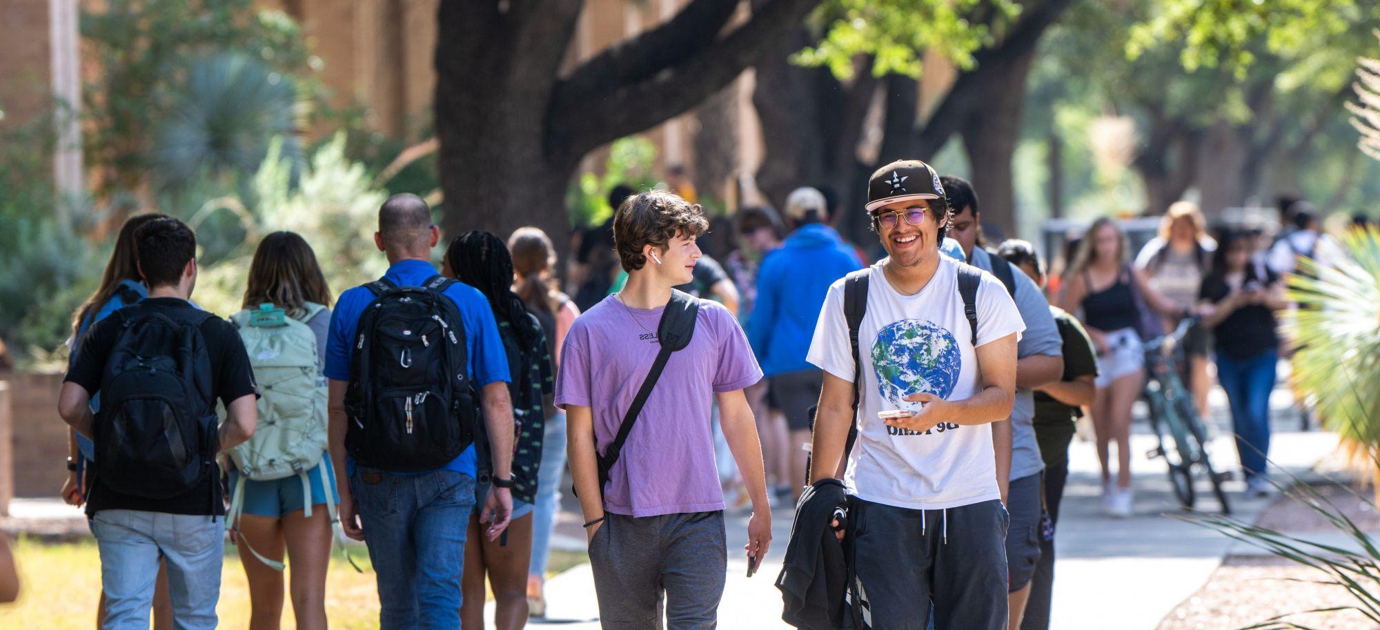 Two male 学生s share a laugh as they walk on a long sidewalk on their first day of school