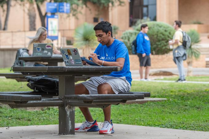 一个学生在外面的桌子上用笔记本电脑学习，另一个学生在学习，两个学生在后面说话.